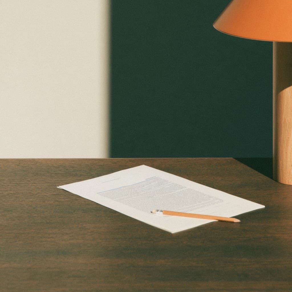 A printed page of text and a pencil resting on a wood desk beside a terracotta lamp.