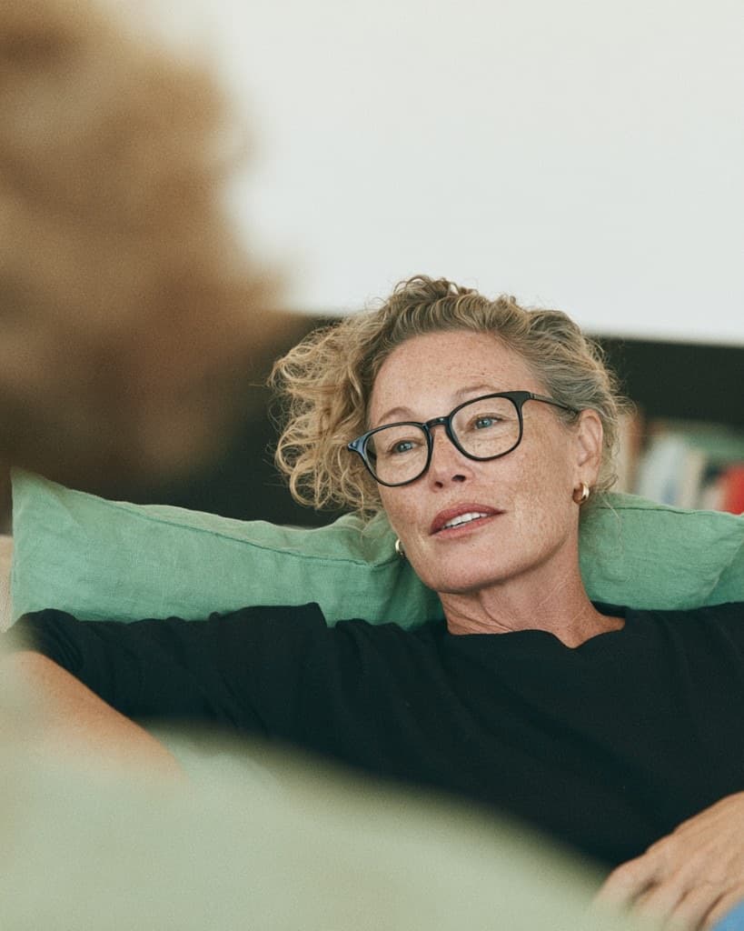 A woman sitting back on a couch in glasses, mid-thought, listening to someone off-frame.
