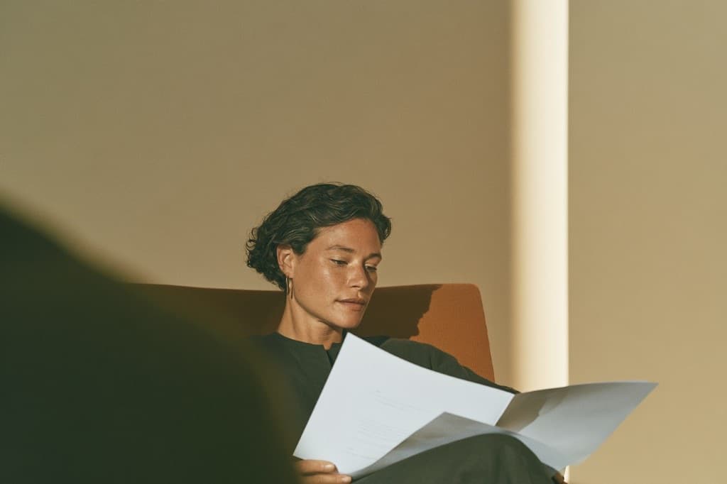 A person sitting in an armchair reading a printed multi-page document in warm afternoon light.