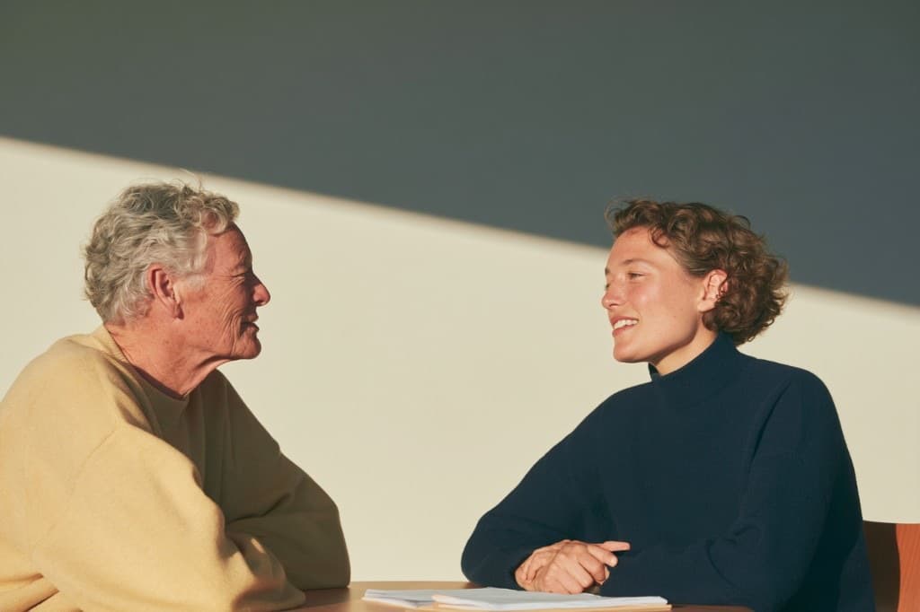 Two people at a table in low sunlight, turned toward each other mid-conversation, notes open in front of them.